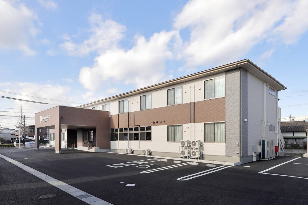 介護施設の建物外観。二階建てモダンデザイン、広々とした駐車場を備える。