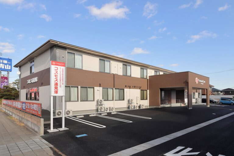 介護施設の建物外観。2階建てのクリーム色と茶色の外壁で、駐車場と看板が見える。
